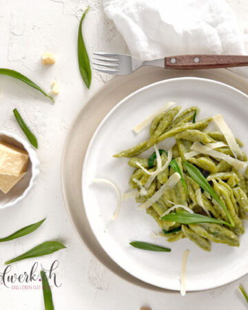 Einfache Bärlauch Scherennudeln auf weissem Teller angerichtet. Daneben eine Schale mit Käse, weisse Serviette und eine Gabel. Um den Teller liegen einige Bärlauchblätter